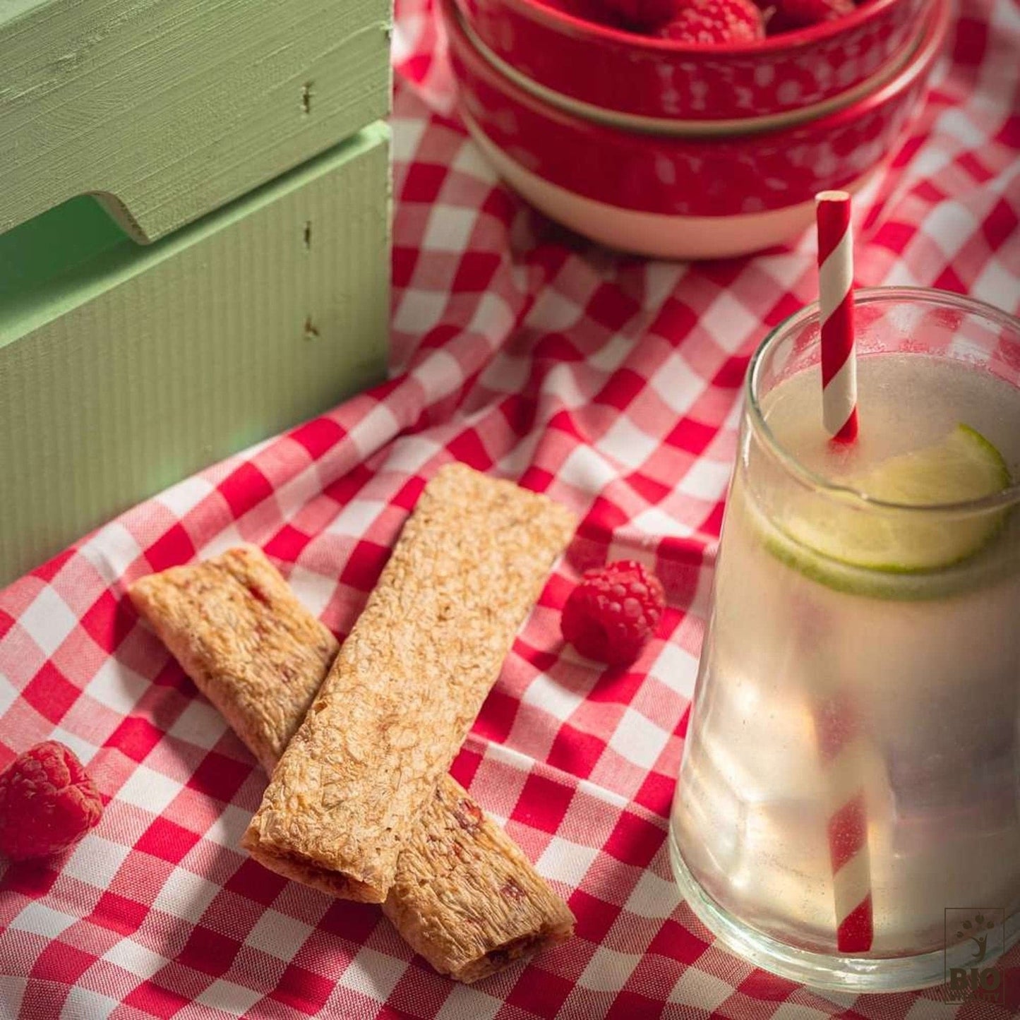 Une mise en place pique-nique avec des barres granola vegan Goûtine framboise boîte Bio - 125g dAGLINA sur une nappe à carreaux rouges, accompagnées dun verre deau infusée au citron vert et dun bol de framboises, vous permettant de savourer les gourmandises sans gluten dans létreinte sereine de la nature.
