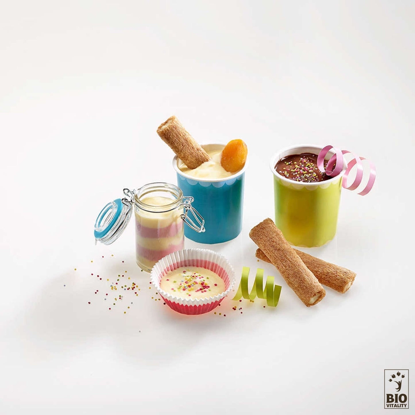 Desserts variés dans des coupes colorées avec des bâtonnets de biscuits et des paillettes, ainsi quune mousse à étages dans un pot. À proximité, un ruban présente la Goûtine noix de coco Bio dAGLINA - 125 g, élaborée à partir de farine de sarrasin pour une délicieuse friandise sans gluten.