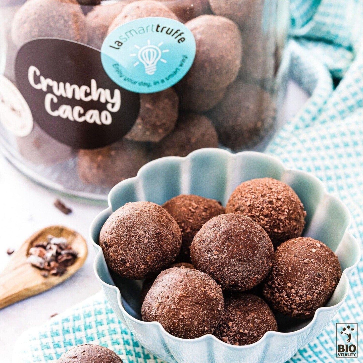 Un bol bleu rempli de truffes au chocolat est le point central, avec un pot de LE SMART CAKE Smart Truffes Crunchy Cacao - 500g en arrière-plan. Une cuillère en bois avec des éclats de cacao sur un tissu à motifs complète cette exquise composition de truffes sans gluten.