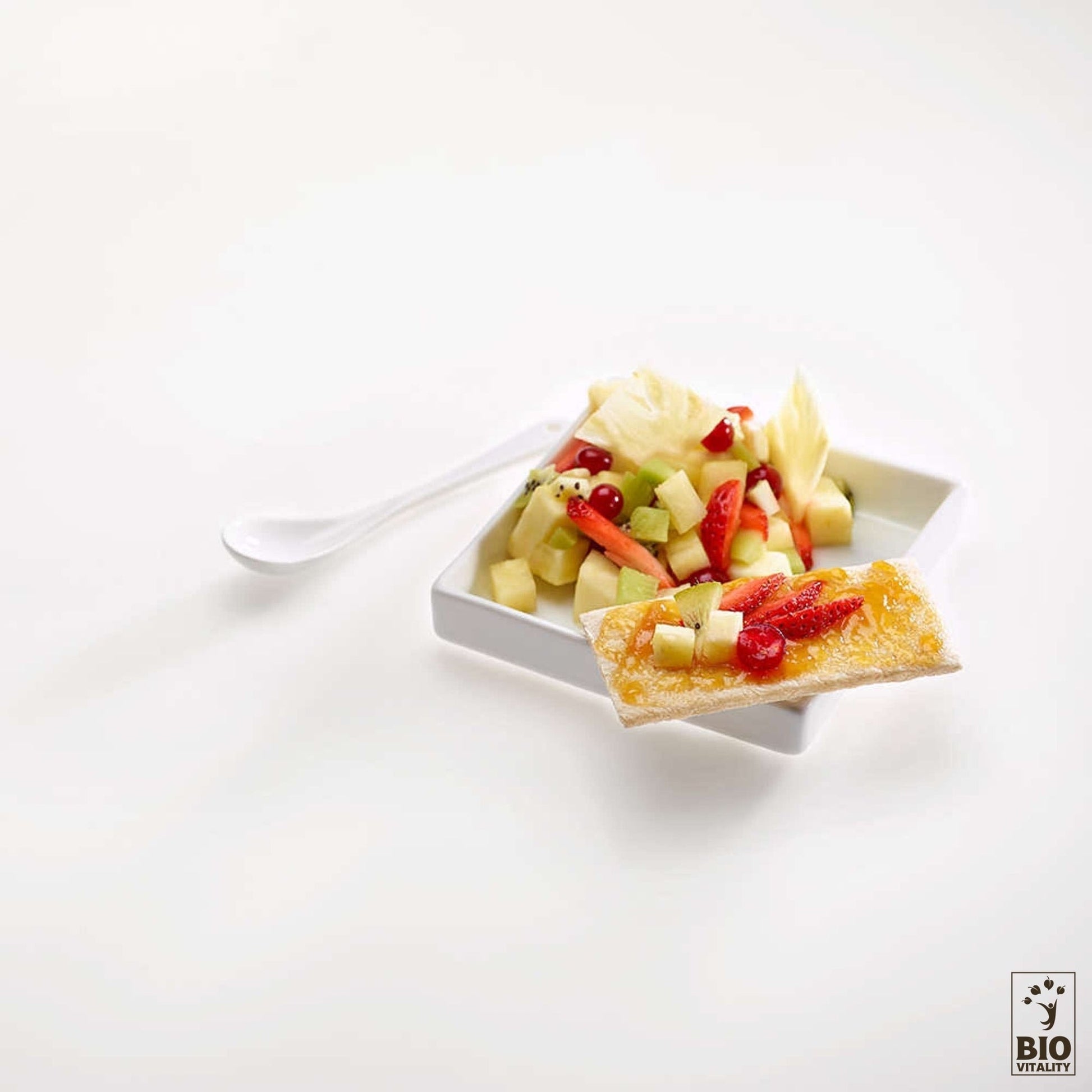 Une assiette carrée blanche avec une salade de fruits rafraîchissante composée de kiwi, de pastèque, dananas et de baies est posée de manière alléchante à côté dune boîte de 5 céréales AGLINA Galette Bio. Une cuillère blanche repose à proximité, prête à déguster ce délice sans gluten.