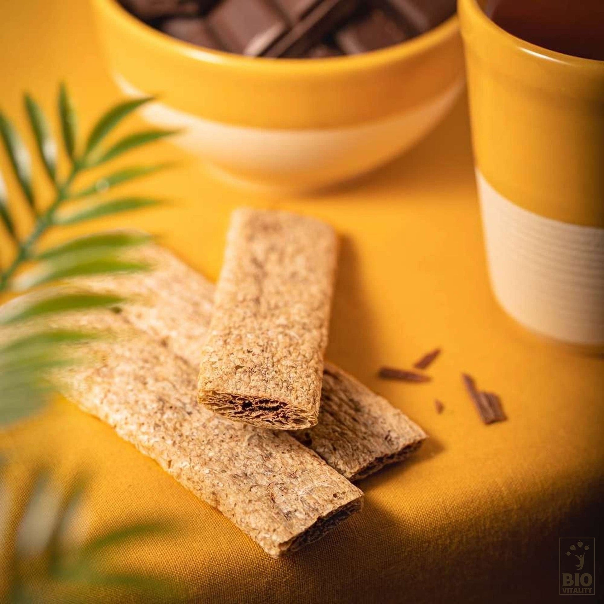 Deux pains plats croustillants sur une surface jaune, une tasse de boisson brune et la boîte de chocolat noir Goûtine dAGLINA Bio - 125g en arrière-plan, partiellement cachée par des feuilles vertes. Savourez cette gourmandise végétalienne saine, sans gluten.