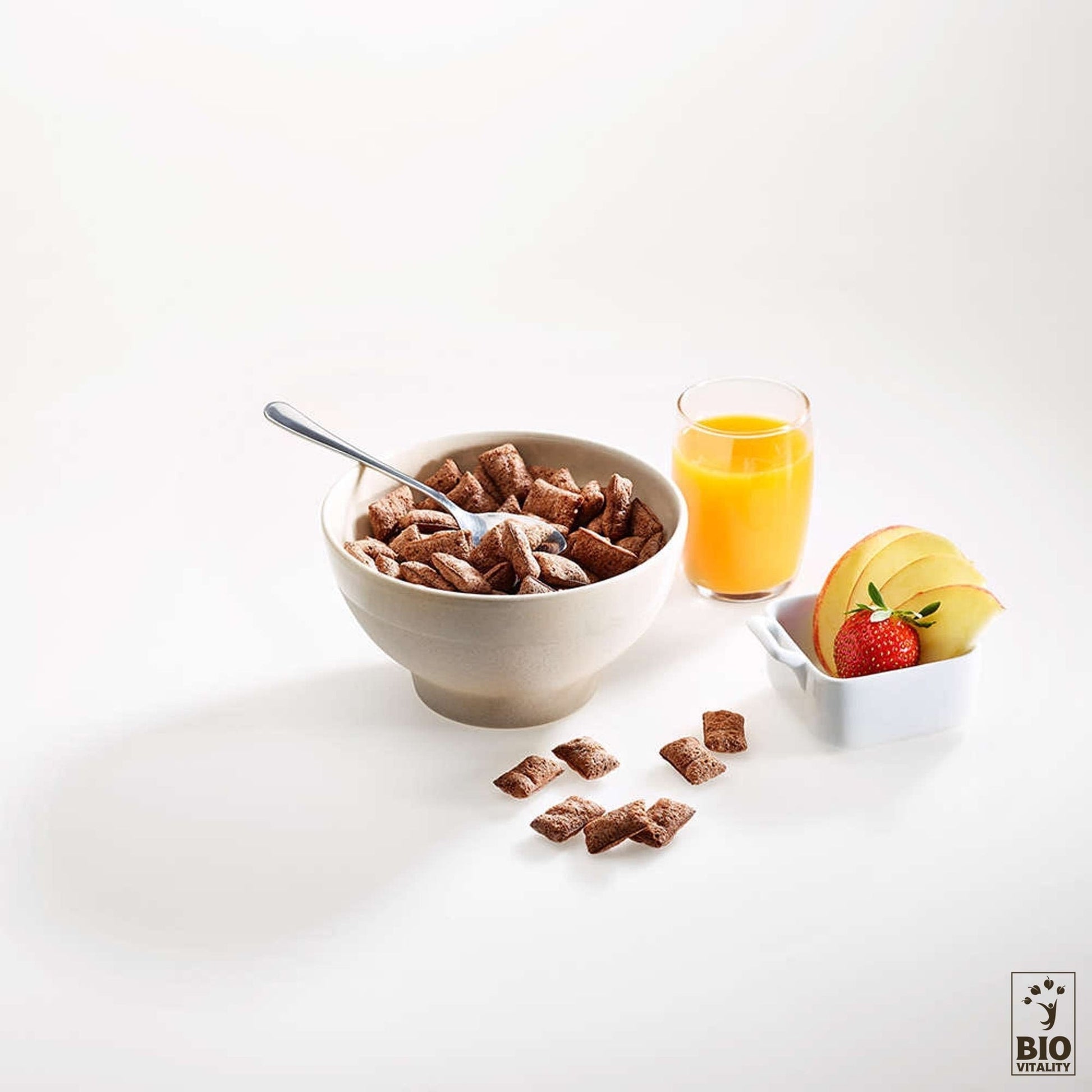 Sur une surface blanche, un bol de céréales Ptit Carré cacao & noisettes Bio dAGLINA est entouré dune cuillère, de morceaux de céréales éparpillés, de jus dorange et de tranches de pomme avec une fraise. Choix de petit-déjeuner parfait sans gluten et vegan friendly.
