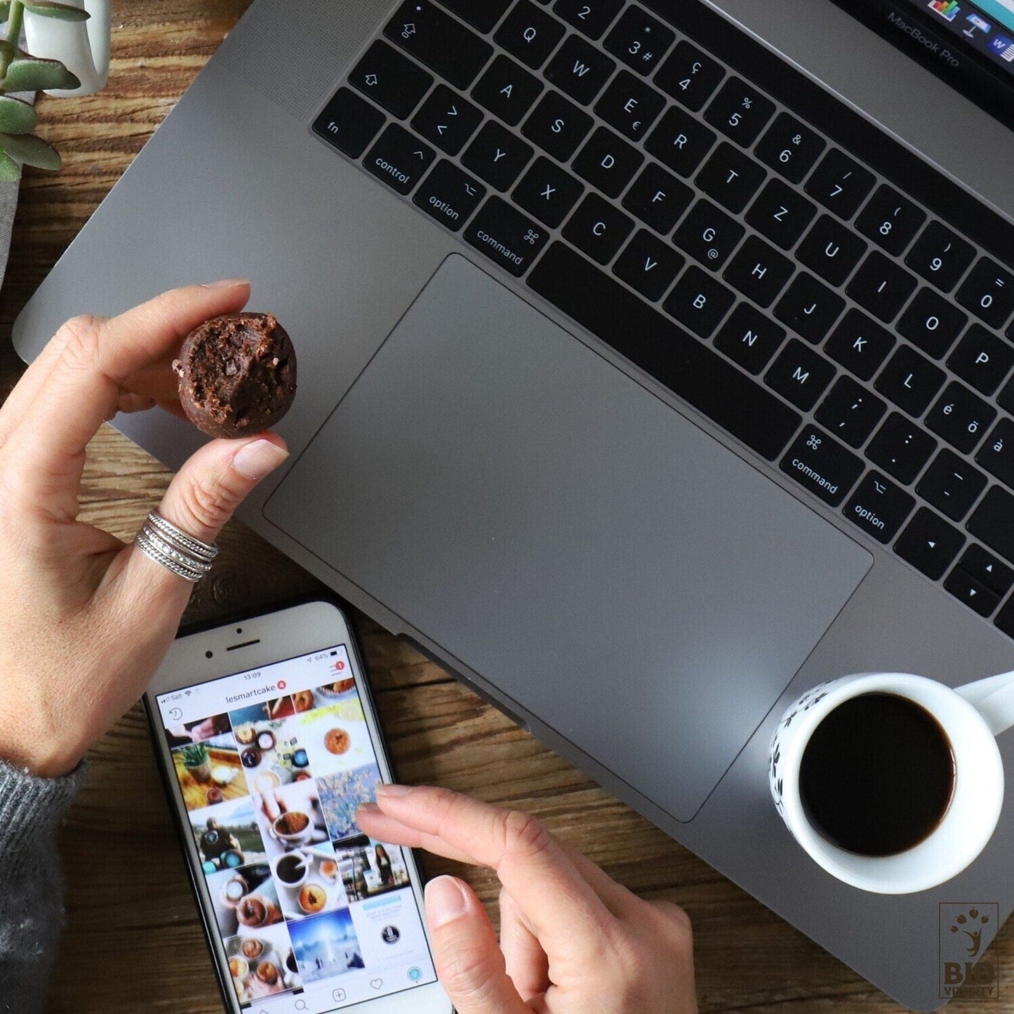 Une personne tient un muffin au chocolat et un smartphone affichant Instagram, à côté dun ordinateur portable et dun café sur une table en bois. À proximité, les Smart Truffes Crunchy Cacao - 2 pièces de LE SMART CAKE ajoutent une touche gourmande à la scène chaleureuse.
