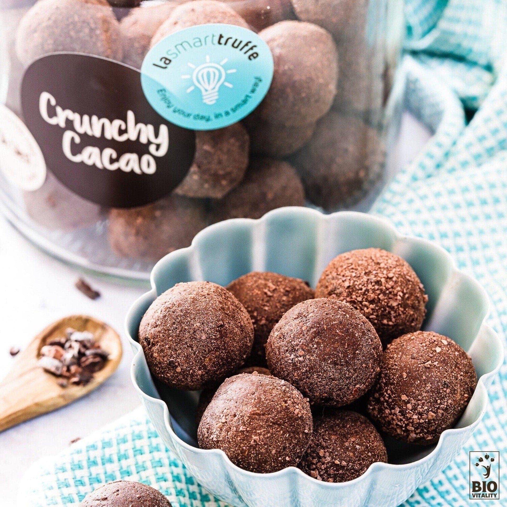 Un bol bleu rempli de truffes au chocolat est le point central, avec un pot de LE SMART CAKE Smart Truffes Crunchy Cacao - 500g en arrière-plan. Une cuillère en bois avec des éclats de cacao sur un tissu à motifs complète cette exquise composition de truffes sans gluten.