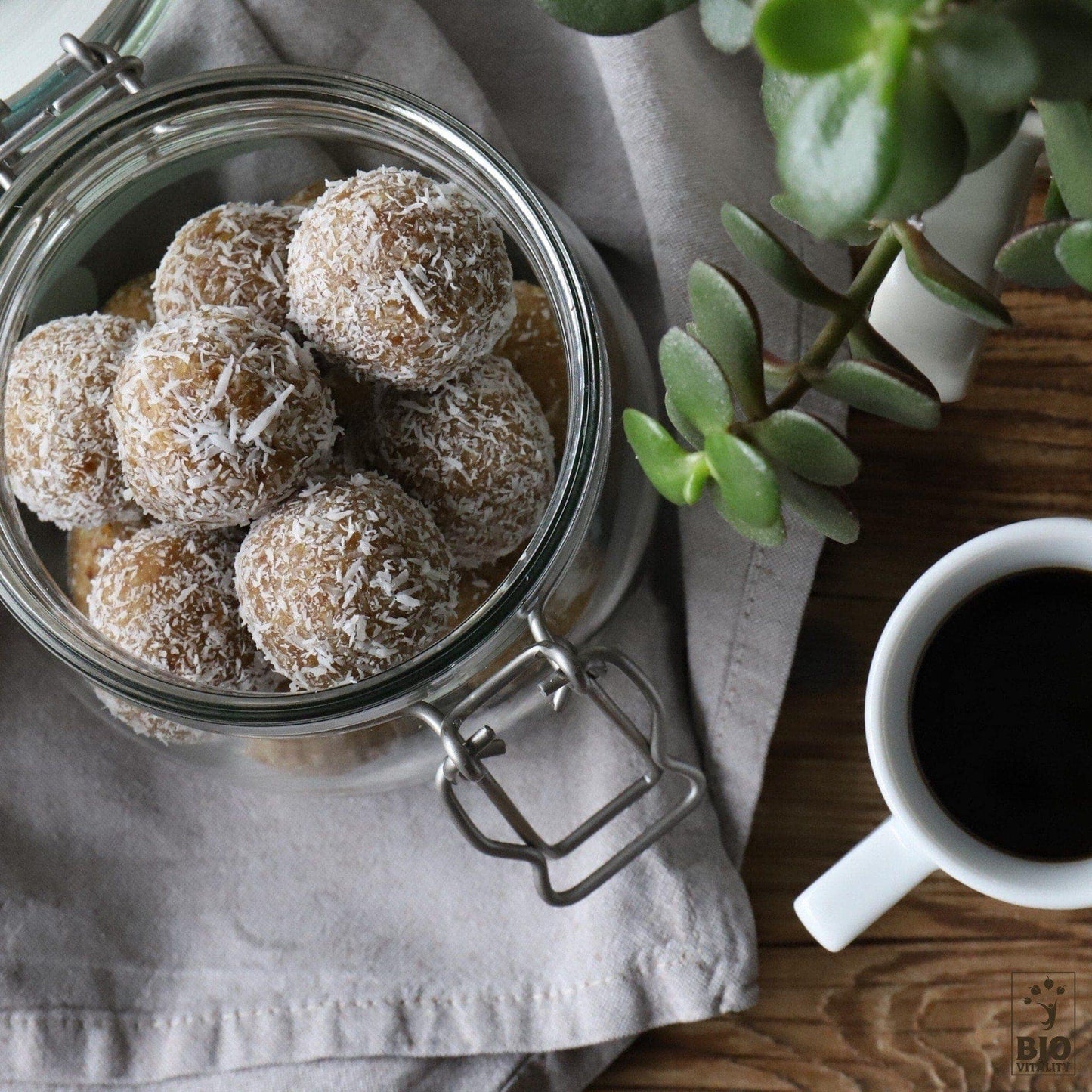 Un pot en verre de 500 g de Truffes Noix de Coco Smart de LE SMART CAKE repose sur une table en bois. À côté, une tasse de café noir complète la scène avec une plante verte vivace. Ces friandises sans gluten sont parfaites pour votre prochain regain dénergie.