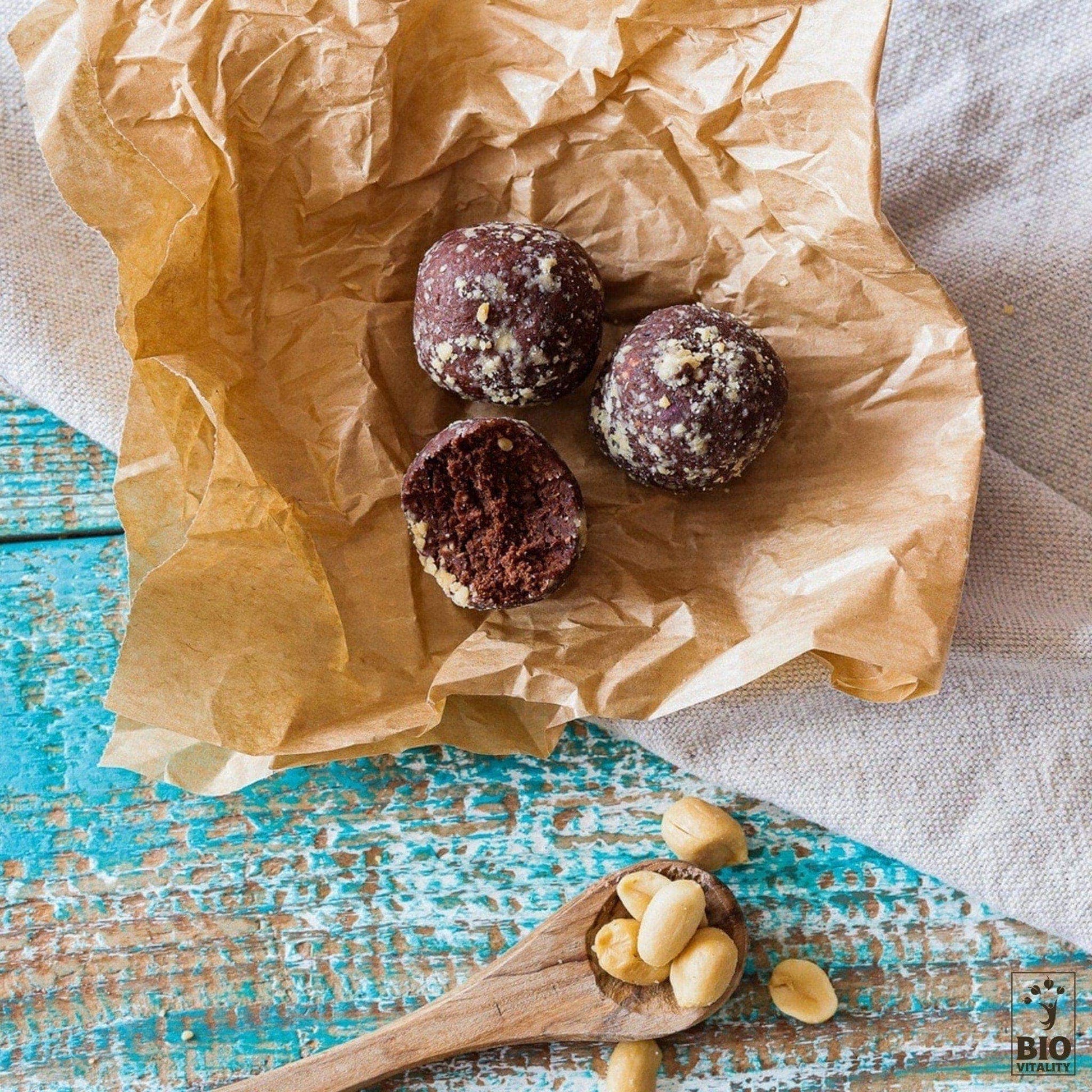 Deux Smart Truffes Peanut Cacao de LE SMART CAKE, dont une partiellement mangée, reposent sur du papier brun froissé à côté dune cuillère en bois de noix sur une surface en bois bleu, incarnant une gourmandise réinventée avec des ingrédients de qualité supérieure.